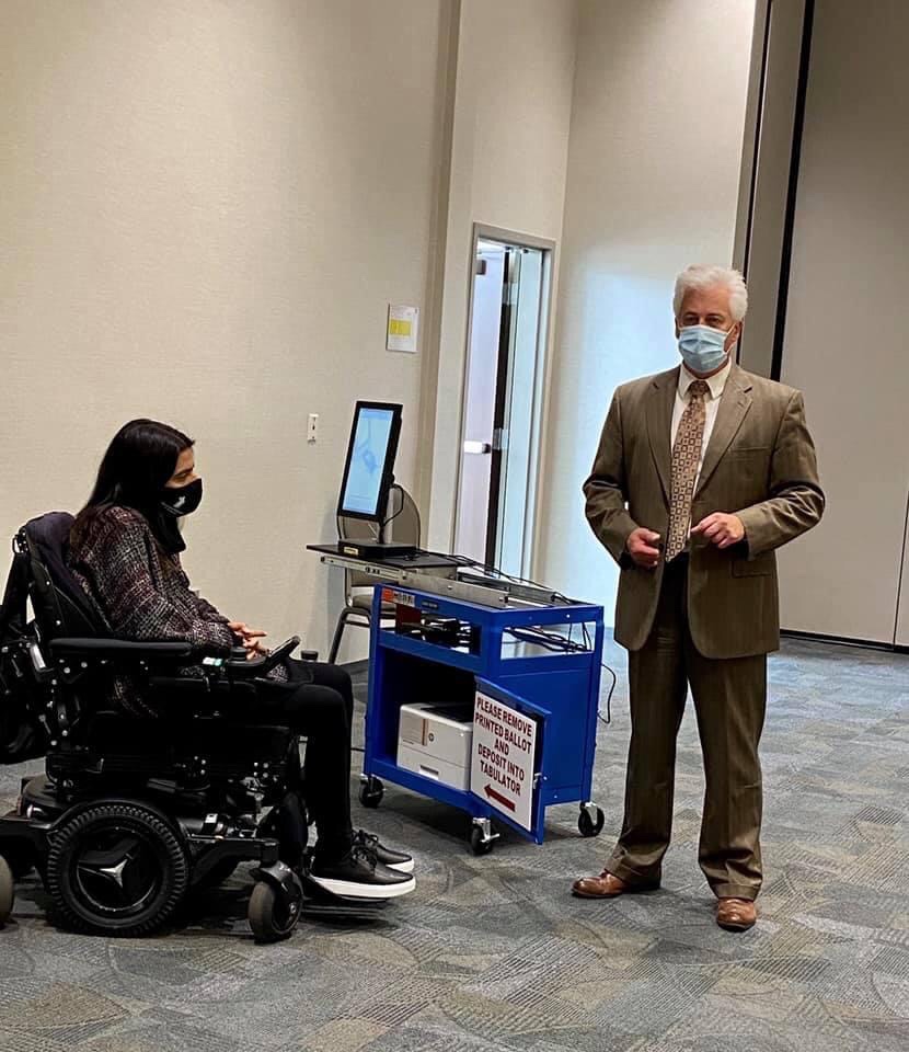 A woman sits in her power wheelchair in front of a touch screen used for accessible voting for those with disabilities. A man stands beside her showing her how to use it.