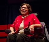 Picture of Judy at her TED talk. She is smiling at the camera, sitting in her motorized wheelchair.